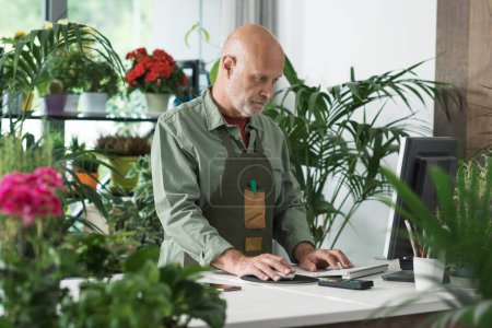 Photo for Professional florist working in his plant shop, he is using a computer and taking orders online - Royalty Free Image