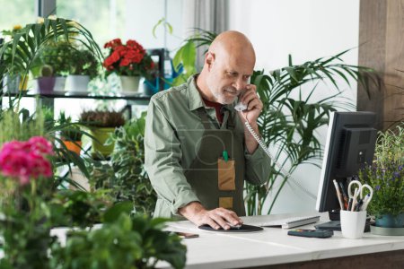 Téléchargez les photos : Fleuriste professionnel travaillant dans son atelier, il prend des commandes au téléphone et utilise un ordinateur - en image libre de droit