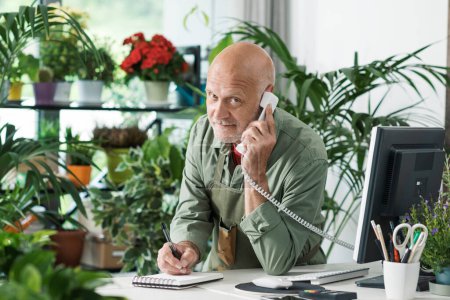Foto de Florista profesional que trabaja en su tienda de plantas, está hablando por teléfono y tomando pedidos, concepto de pequeña empresa - Imagen libre de derechos