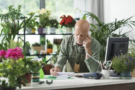 Photo for Business owner working in his flower shop, he is checking an invoice and having a phone call - Royalty Free Image
