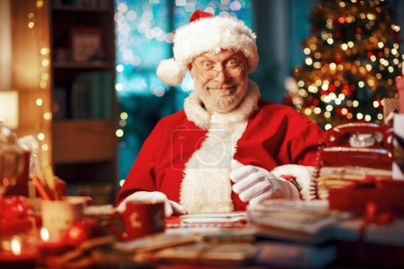 Téléchargez les photos : Joyeux Père Noël assis à son bureau et souriant à la caméra, Noël et vacances concept - en image libre de droit