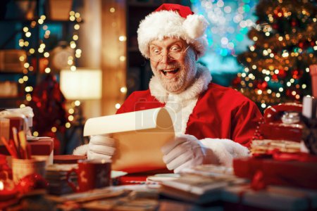 Téléchargez les photos : Joyeux Père Noël joyeux assis à son bureau et lire des lettres, Noël et vacances concept - en image libre de droit