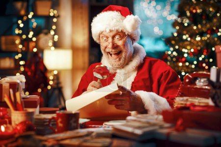 Téléchargez les photos : Joyeux Père Noël joyeux assis à son bureau et lisant des lettres à l'aide d'une loupe, Noël et vacances concept - en image libre de droit