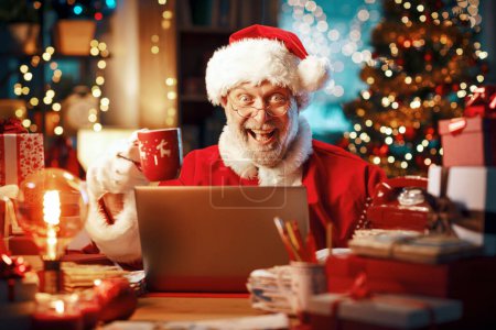 Téléchargez les photos : Joyeux Père Noël assis à son bureau et faisant une pause café, il regarde des vidéos en ligne sur son ordinateur portable - en image libre de droit