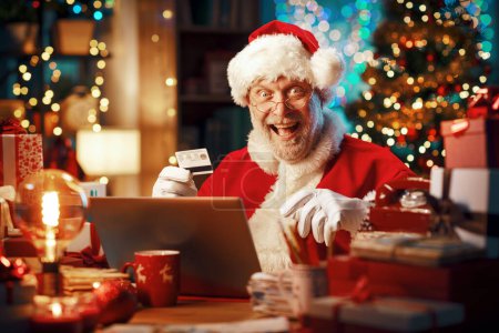 Téléchargez les photos : Père Noël assis à son bureau et faisant des achats en ligne pour Noël avec une carte de crédit, il est joyeux et souriant - en image libre de droit