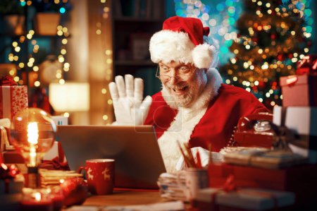 Téléchargez les photos : Joyeux Père Noël ayant un appel vidéo avec son ordinateur portable, il fait signe à l'écran de l'ordinateur et souhaite un joyeux Noël - en image libre de droit