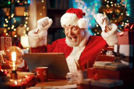 Téléchargez les photos : Joyeux Père Noël assis à son bureau et se connectant en ligne, il a reçu une belle surprise en ligne et il célèbre - en image libre de droit