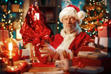 Téléchargez les photos : Père Noël drôle assis à son bureau à la maison et tenant un oeuf de Pâques en chocolat enveloppé, il est en colère et déçu - en image libre de droit
