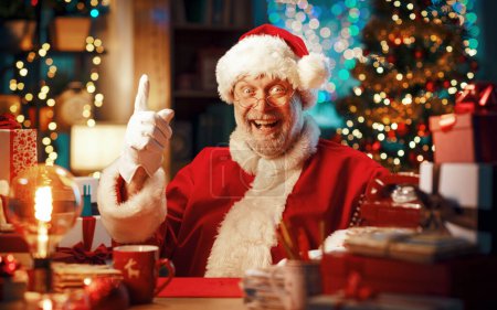 Téléchargez les photos : Joyeux Père Noël assis à son bureau et souriant à la caméra, Noël et vacances concept - en image libre de droit