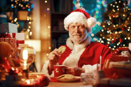 Foto de Feliz Santa Claus sonriente sentado en su escritorio en casa y tomando un descanso para tomar un café, está comiendo galletas y mirando a la cámara - Imagen libre de derechos