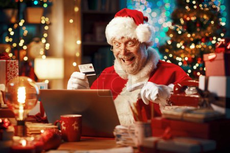 Photo for Santa Claus sitting at his desk and doing online shopping for Christmas using a credit card, he is cheerful and smiling - Royalty Free Image