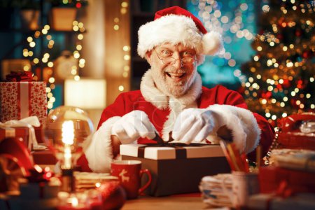 Téléchargez les photos : Joyeux Père Noël assis à son bureau et préparant des cadeaux pour Noël - en image libre de droit