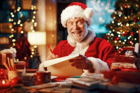 Téléchargez les photos : Joyeux Père Noël joyeux assis à son bureau et écrivant une lettre, Noël et vacances concept - en image libre de droit