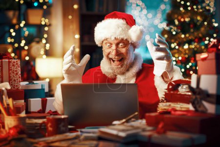 Téléchargez les photos : Joyeux Père Noël assis à son bureau et se connectant en ligne, il a reçu une belle surprise en ligne et il célèbre - en image libre de droit