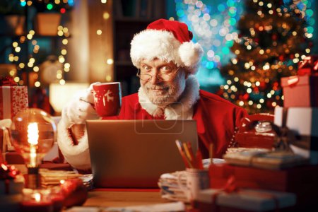 Téléchargez les photos : Joyeux Père Noël assis à son bureau et faisant une pause café, il regarde des vidéos en ligne sur son ordinateur portable - en image libre de droit