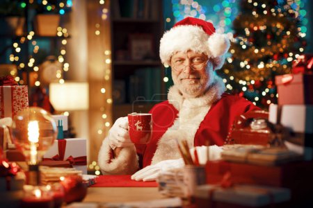 Téléchargez les photos : Joyeux Père Noël souriant assis à son bureau à la maison et ayant une pause café, Noël et vacances concept - en image libre de droit