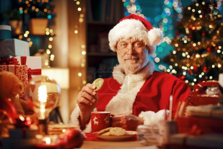 Foto de Feliz Santa Claus sonriente sentado en su escritorio en casa y tomando un descanso para tomar un café, está comiendo galletas y mirando a la cámara - Imagen libre de derechos