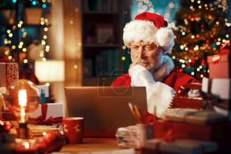 Téléchargez les photos : Père Noël se connecter avec son ordinateur portable à la maison et de penser avec la main sur le menton, Noël et concept de technologie - en image libre de droit