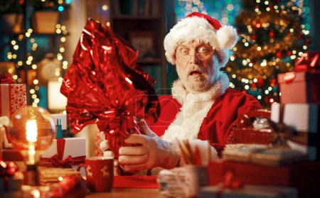 Téléchargez les photos : Drôle Père Noël assis à son bureau à la maison et tenant un oeuf de Pâques en chocolat enveloppé, il est choqué et fixe la caméra avec la bouche ouverte - en image libre de droit