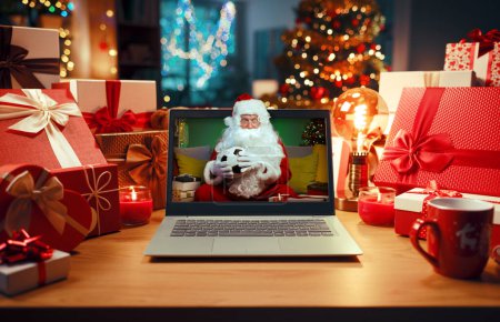 Téléchargez les photos : Joyeux Père Noël regardant le match de football sur un écran d'ordinateur portable entouré de décorations et de cadeaux de Noël, de vacances et de concept sportif - en image libre de droit
