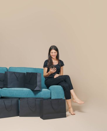 Téléchargez les photos : Femme heureuse assise sur un canapé et faisant des achats en ligne sur Black Friday en utilisant son smartphone - en image libre de droit