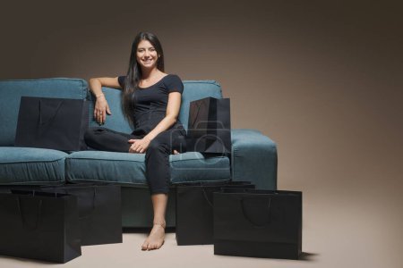 Téléchargez les photos : Femme heureuse assise sur un canapé entouré de sacs à provisions noirs : Vente Black Friday - en image libre de droit