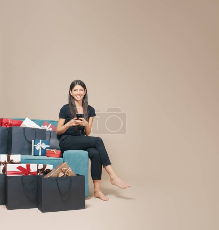 Téléchargez les photos : Femme assise sur le canapé et faisant des achats en ligne sur son smartphone, elle est entourée de cadeaux et de sacs à provisions - en image libre de droit