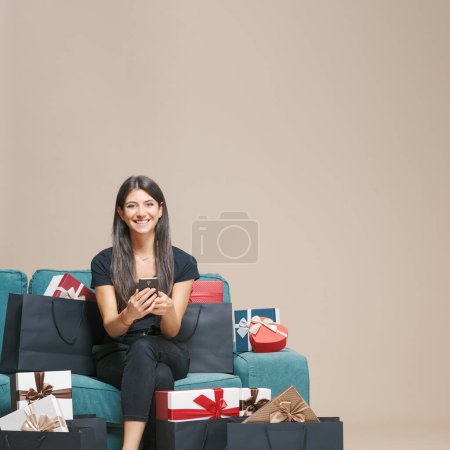 Téléchargez les photos : Femme assise sur le canapé et faisant des achats en ligne sur son smartphone, elle est entourée de cadeaux et de sacs à provisions - en image libre de droit