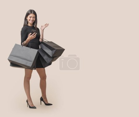 Téléchargez les photos : Belle femme élégante tenant des sacs à provisions et faisant des achats en ligne sur son smartphone, Black Friday vente - en image libre de droit