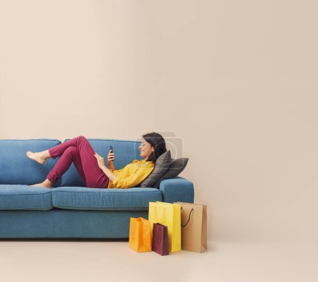 Foto de Mujer joven feliz acostada en el sofá con bolsas de compras y haciendo compras en línea en su teléfono inteligente - Imagen libre de derechos