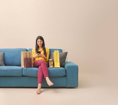 Téléchargez les photos : Joyeux jeune femme assise sur le canapé avec des sacs à provisions et faire des achats en ligne sur son smartphone - en image libre de droit