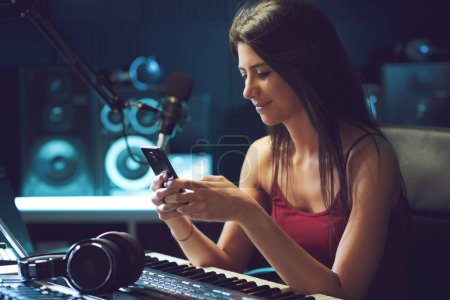 Téléchargez les photos : Jeune artiste travaillant au studio d'enregistrement, elle utilise un smartphone et sourit - en image libre de droit