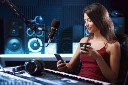 Téléchargez les photos : Jeune artiste travaillant au studio d'enregistrement, elle utilise un smartphone et sourit - en image libre de droit