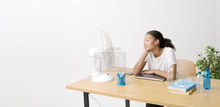 Téléchargez les photos : Jeune femme assise au bureau devant un ventilateur électrique, elle souffre de la chaleur - en image libre de droit