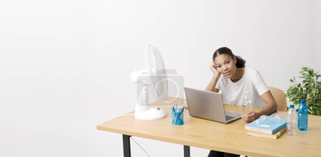 Téléchargez les photos : Jeune femme assise au bureau devant un ventilateur électrique, elle souffre de la chaleur - en image libre de droit