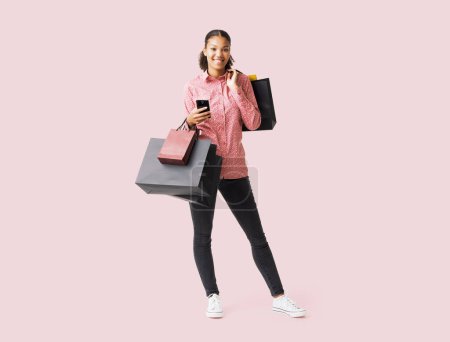 Téléchargez les photos : Joyeux jeune femme faisant du shopping le Black Friday, elle tient son smartphone et de nombreux sacs à provisions - en image libre de droit
