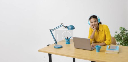 Téléchargez les photos : Jeune femme assise au bureau et regardant des vidéos en ligne sur son ordinateur portable, elle porte un casque - en image libre de droit