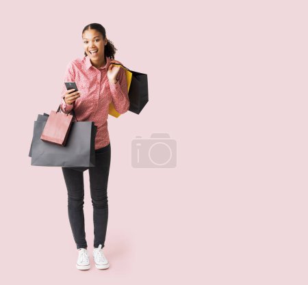 Téléchargez les photos : Joyeux jeune femme faisant du shopping le Black Friday, elle tient son smartphone et de nombreux sacs à provisions - en image libre de droit