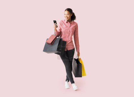 Foto de Mujer joven feliz haciendo compras en Viernes Negro, ella está sosteniendo su teléfono inteligente y muchas bolsas de compras - Imagen libre de derechos