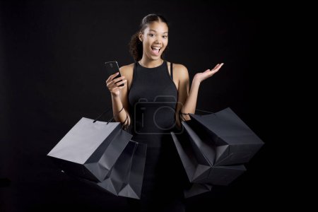 Téléchargez les photos : Belle jeune femme tenant des sacs à provisions noirs et un smartphone : concept de vente Black Friday - en image libre de droit