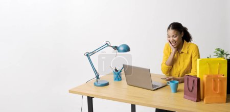 Foto de Mujer joven emocionada sentada en su escritorio y haciendo compras en línea en su computadora portátil, ella está revisando ofertas y descuentos - Imagen libre de derechos