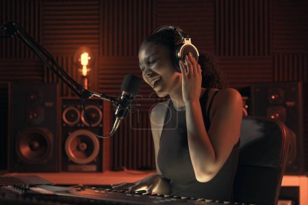Téléchargez les photos : Jeune artiste féminine enregistrant une chanson en studio, elle chante et joue du clavier - en image libre de droit