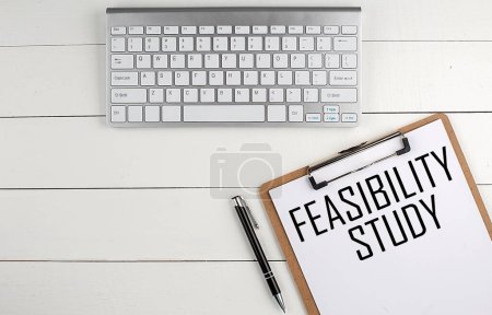 Home office workspace with keyboard, clipboard and pen with text FEASIBILITY STUDY on a white wooden background , business concept