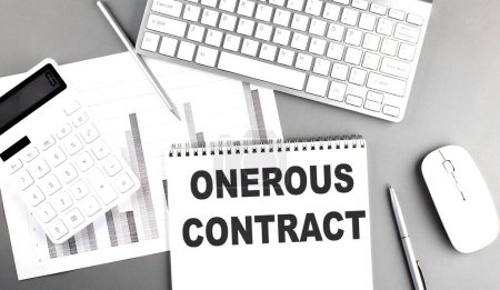 Foto de ONEROUS CONTRACT text written on a notebook on grey background with chart and keyboard, business concept - Imagen libre de derechos