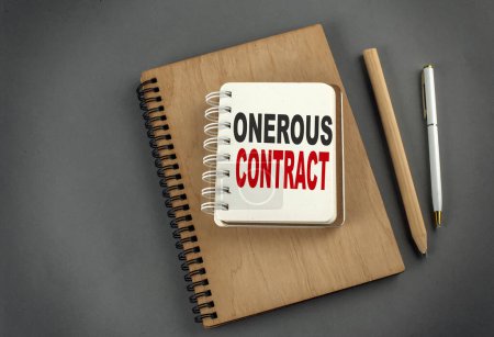 Photo for ONEROUS CONTRACT text on a notebook with pen and pencil on grey background - Royalty Free Image