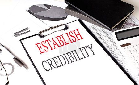 Photo for ESTABLISH CREDIBILITY text on paper clipboard with chart and notebook on a withe background - Royalty Free Image