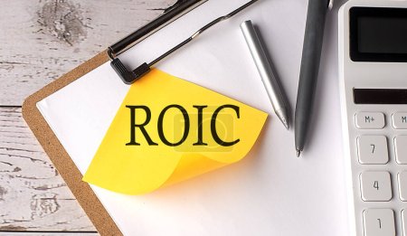Palabra ROIC en un adhesivo amarillo con calculadora, pluma y portapapeles