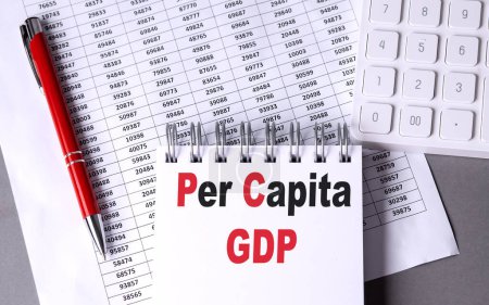 PER CAPITA GDP text on a notebook with pen, calculator and chart on grey background