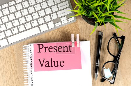 PRESENT VALUE text on a pink sticky on notebook with laptop, pen and glasses. 