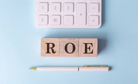 ROE word on a wooden block with pen and calculator on blue background . 